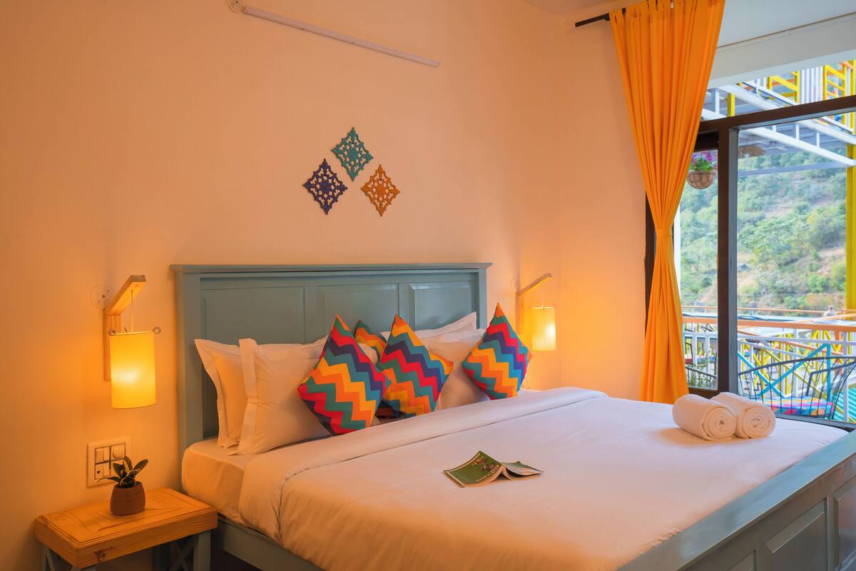 A bed with colorful zigzag pillows, a bedside table with a plant, and a window with a view of greenery.