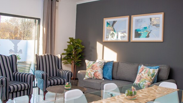 Una sala de estar moderna con sillones a rayas, un sofá gris con cojines coloridos, una mesa de centro de vidrio y dos cuadros abstractos enmarcados en la pared.