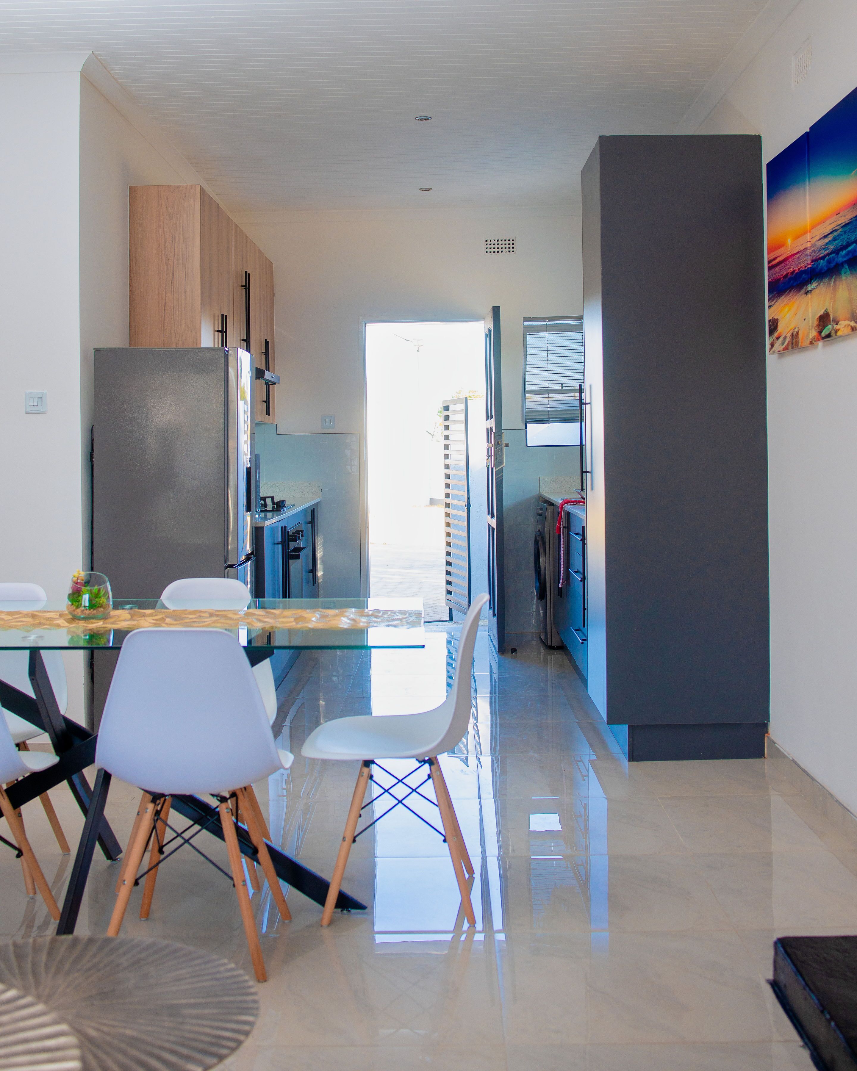 A modern kitchen with a glass dining table, white chairs, and a stainless steel refrigerator.