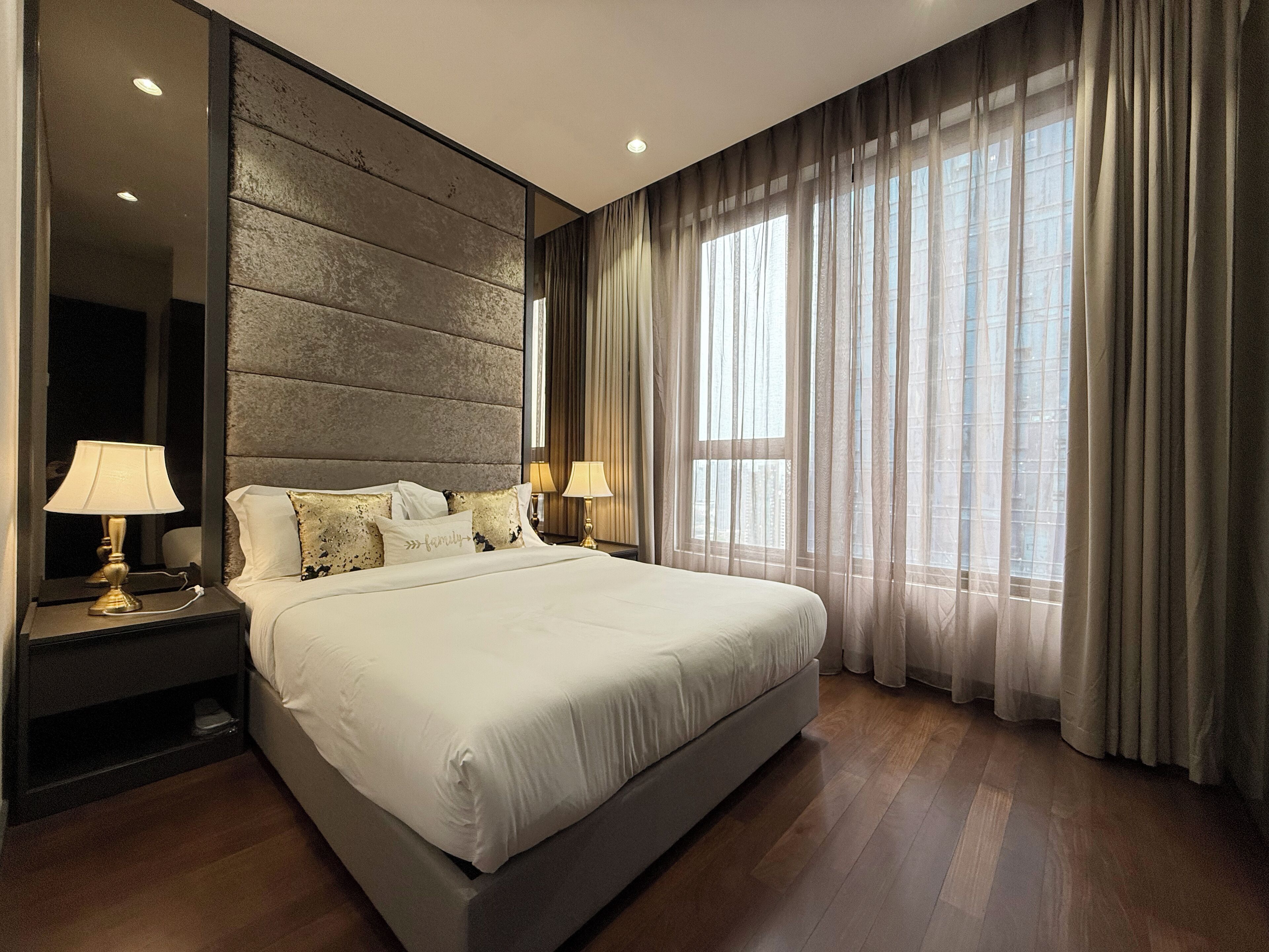 A modern bedroom with a large bed, two bedside tables with lamps, a large mirror, and a view of the city through sheer curtains.