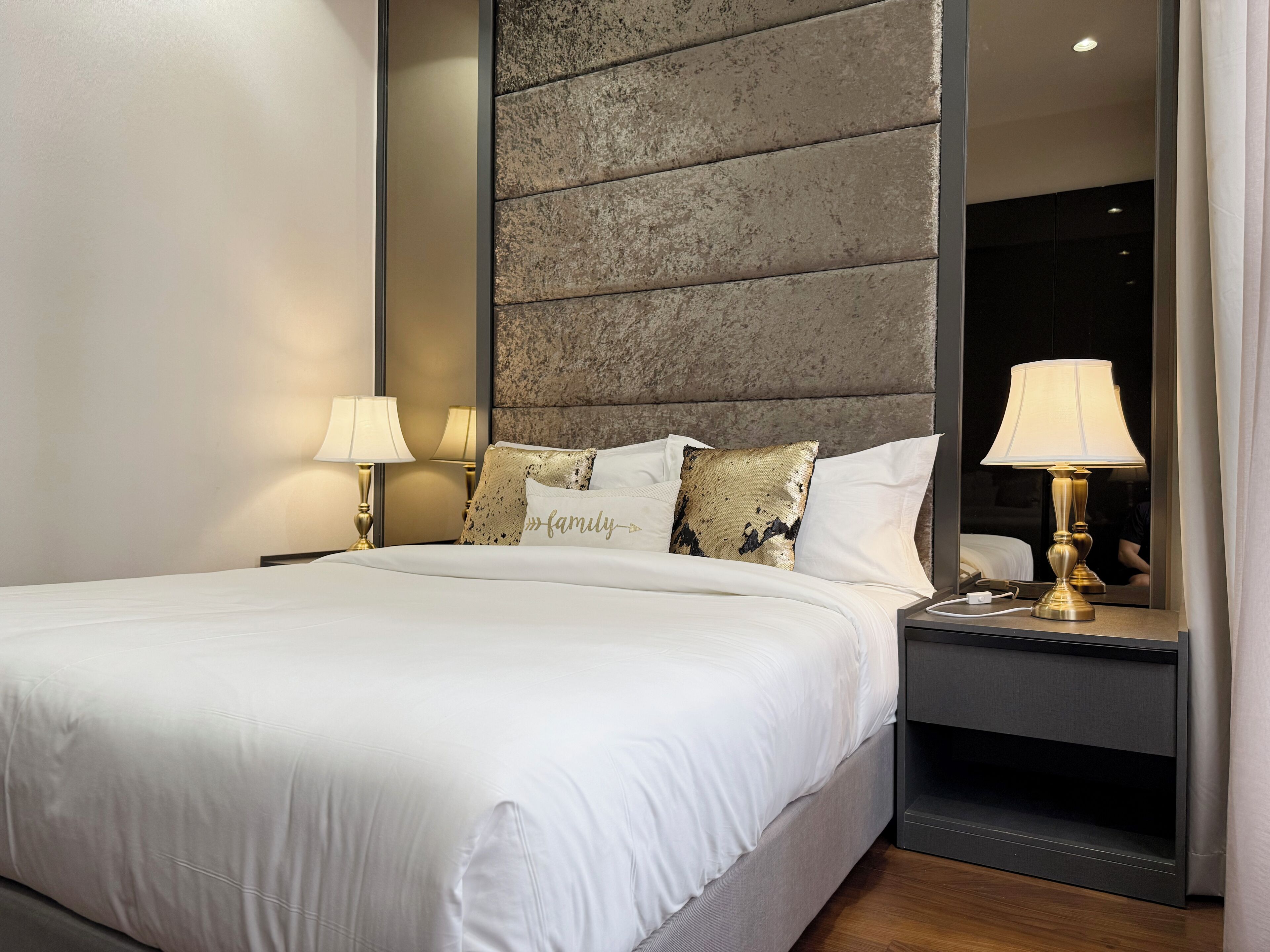 A neatly made bed with white linens, a headboard with a textured stone design, two bedside lamps, and a nightstand with a mirror.