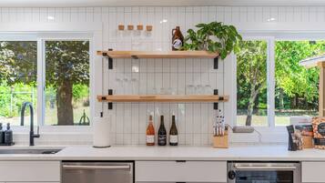 Cocina moderna con armarios blancos, encimera de madera y un amplio ventanal con vistas a árboles. Hay botellas de vino y copas sobre la encimera, y una planta en el alféizar de la ventana.