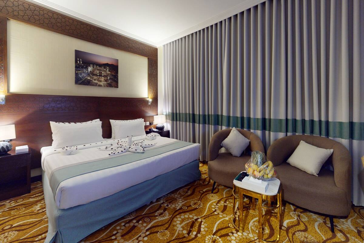 A hotel room with a large bed, a sofa, a small table, and a view of a cityscape through curtains.