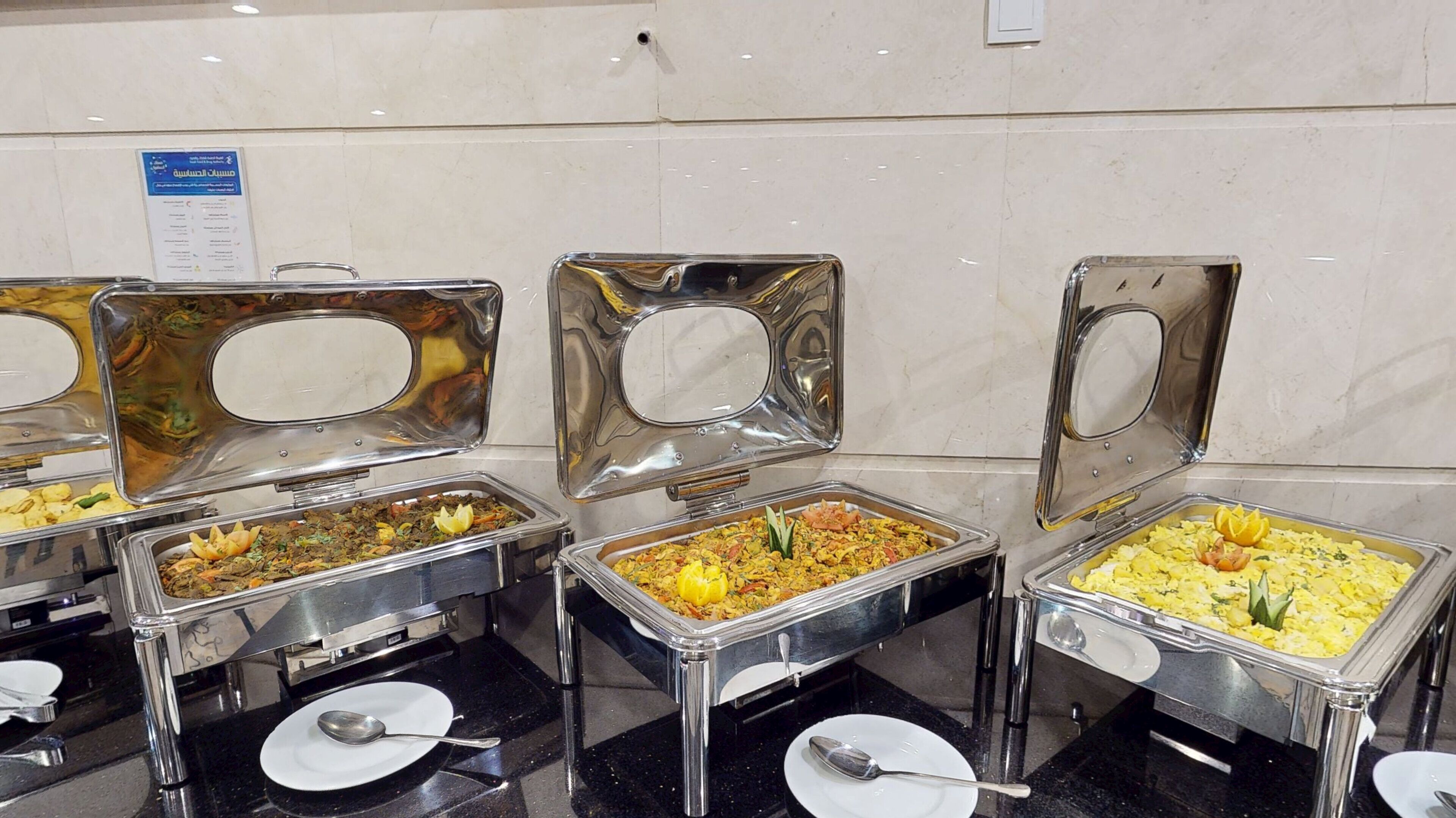 Dispositiu de buffet amb nombroses safates metàl·liques que contenen diferents plats, cadascun amb una cullera i un plat a la barra.