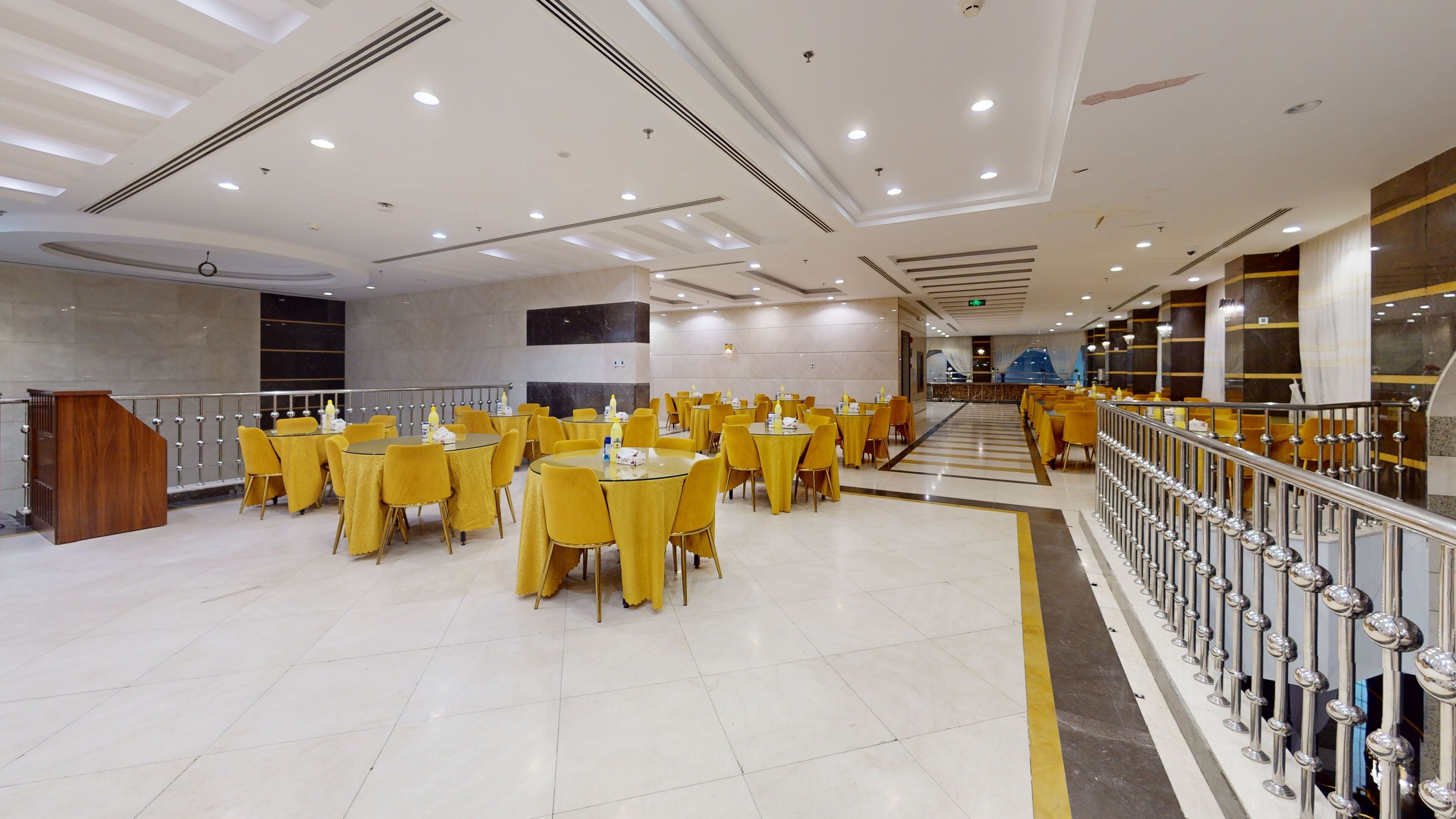A spacious indoor dining area with multiple round tables and yellow chairs, a staircase, and a railing along the upper walkway.