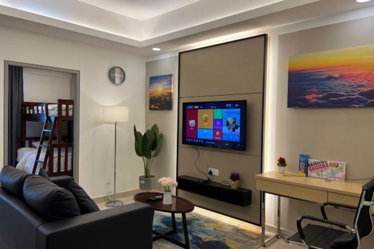 Een moderne hotelkamer met een flatscreen televisie aan de muur, een bureau met een stoel, een bank, een kleine tafel met een plant en een groot raam met uitzicht op de lucht.