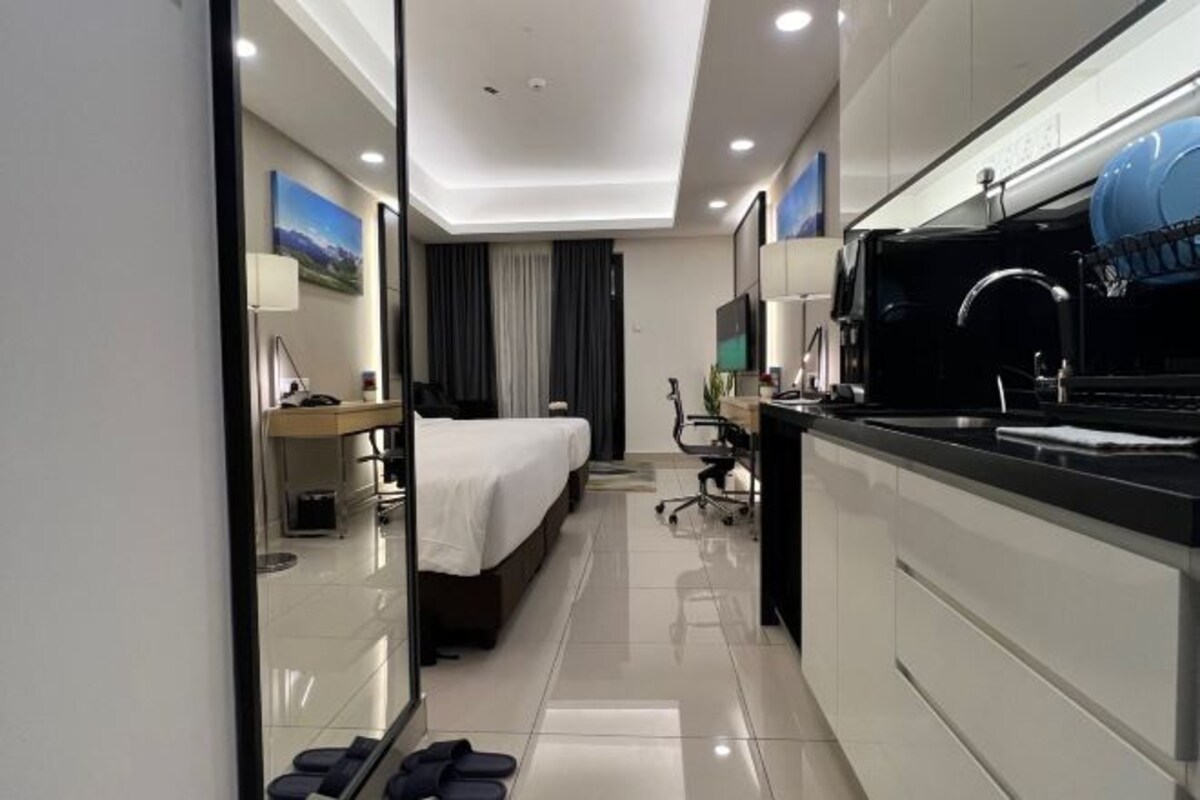 Een moderne hotelkamer met een bed, nachtkastje, lamp en een kitchenette met een spoelbak en kookplaat.