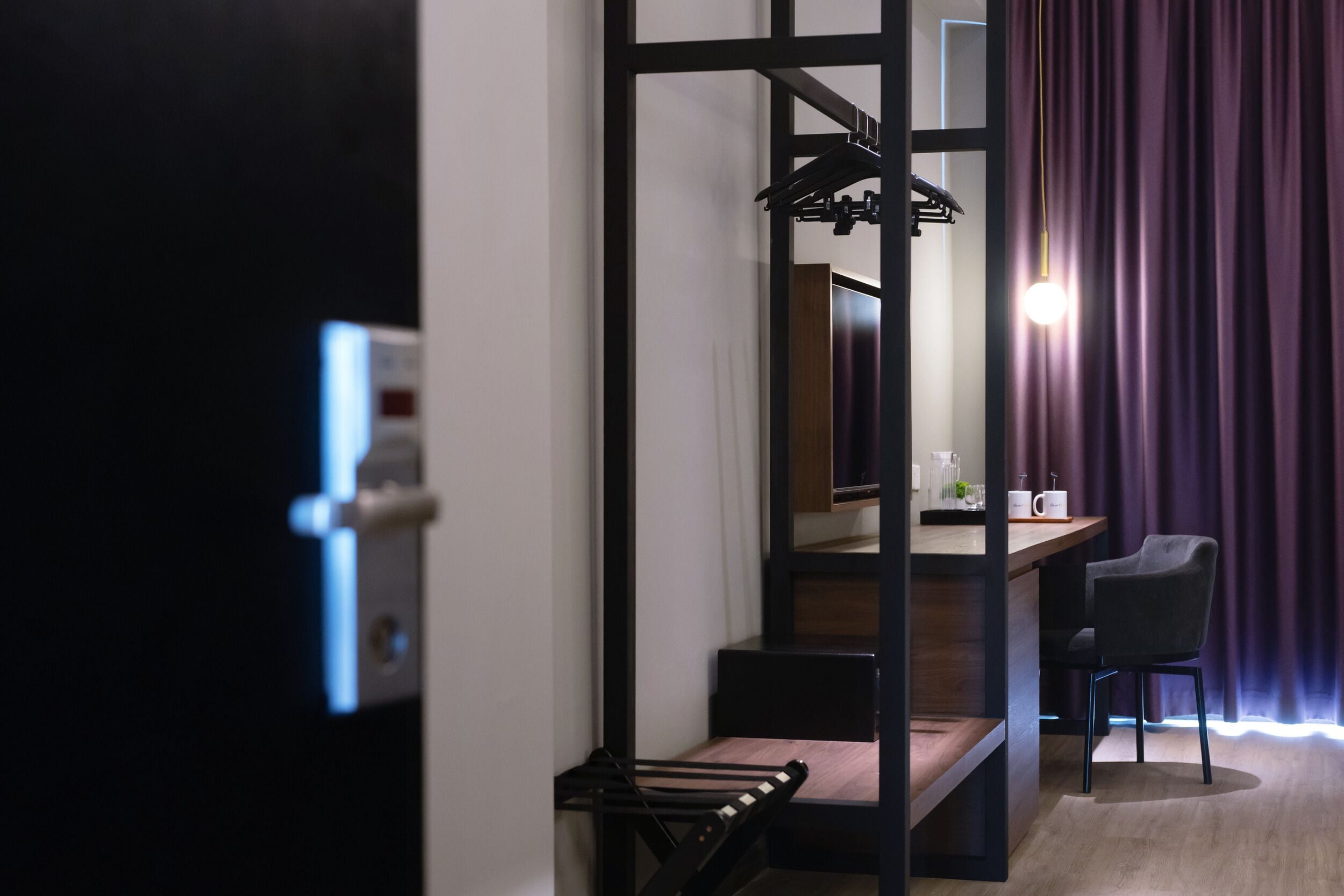 A modern hotel room with a wooden staircase, a desk with a chair, and a view of the outside through a window with purple curtains.