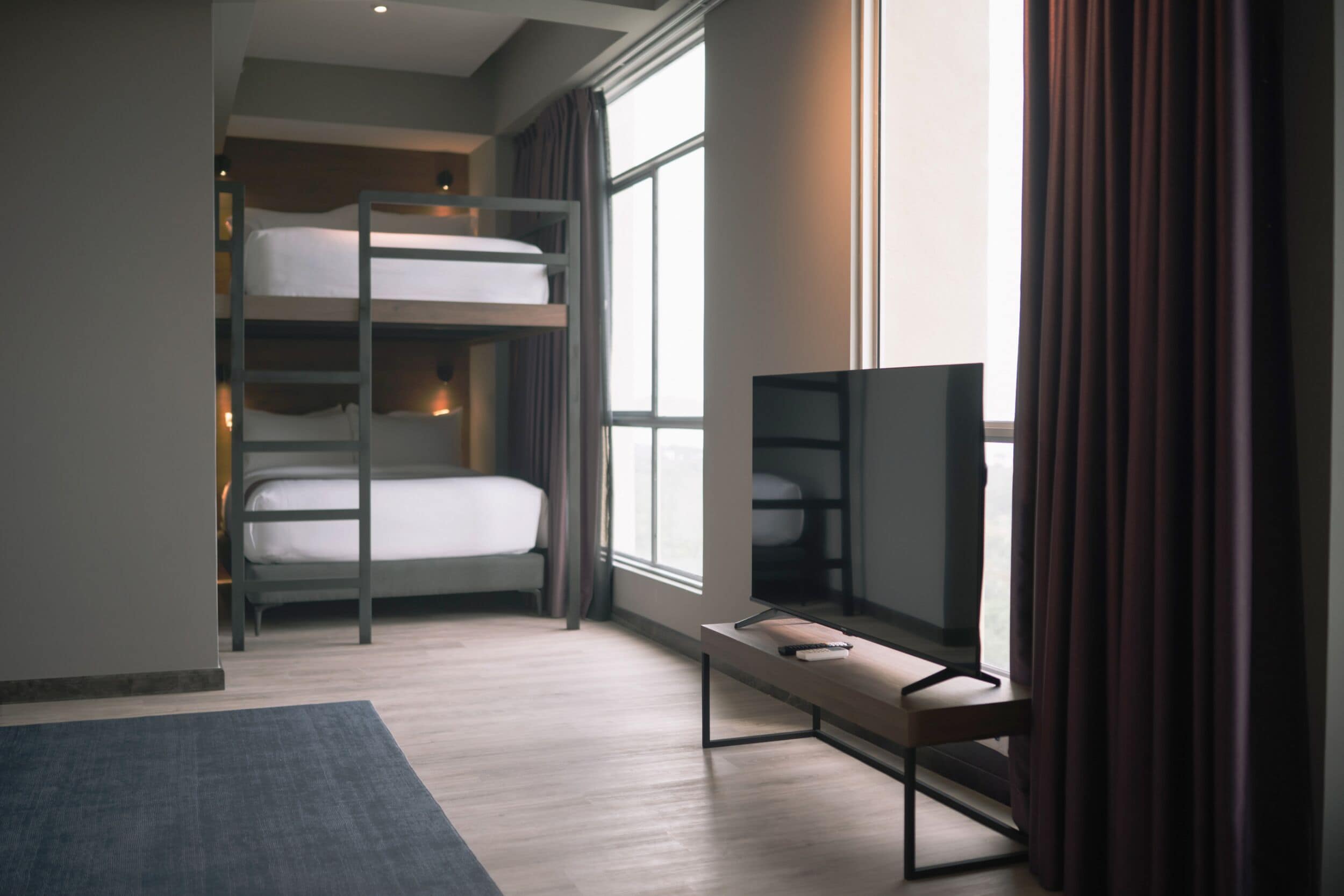 A modern hotel room with bunk beds, a flat-screen TV, and a large window with curtains.