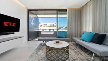 A modern living room with a flat-screen TV, a sofa, a coffee table, and a balcony with a view.