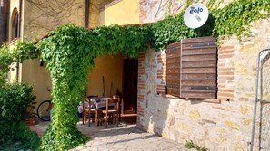Relax in a charming outdoor seating area with a wooden table and chairs under a lush vine canopy, adjacent to a stone wall with a wooden shutter.