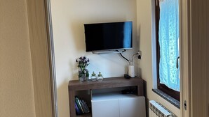 Une pièce de taille modeste avec un plafond en bois, un téléviseur à écran plat fixé au-dessus d’une petite armoire, un radiateur et une fenêtre ornée d’un rideau à motifs.