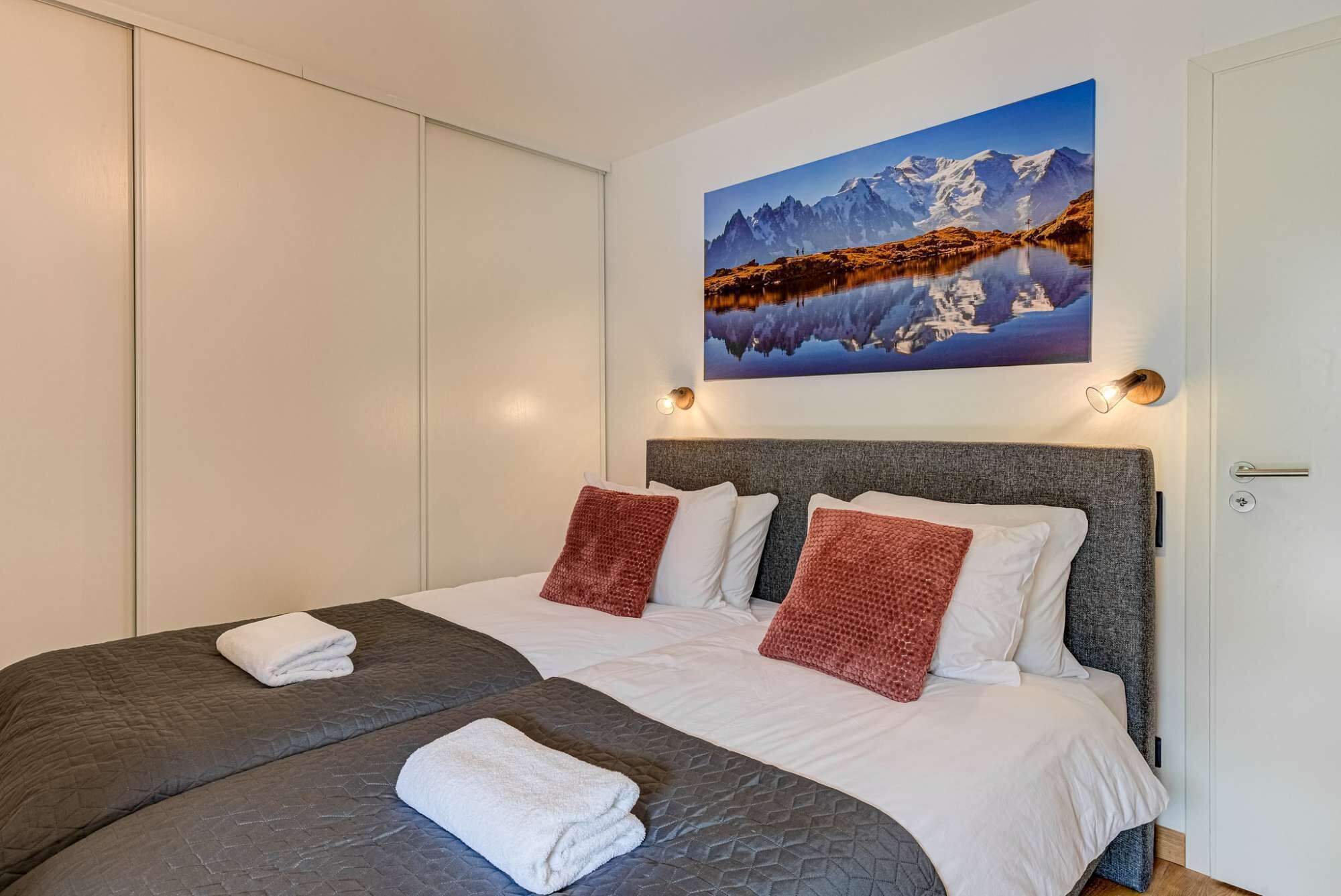 Een tweepersoonsbed met grijs en wit beddengoed, twee rode kussens en een berglandschap schilderij erboven.