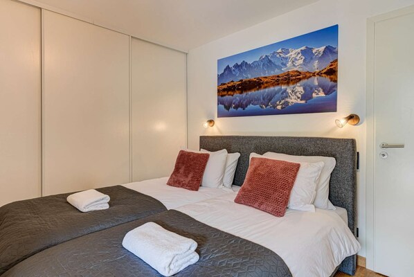 A double bed with grey and white bedding, two red pillows, and a mountain landscape painting above it.