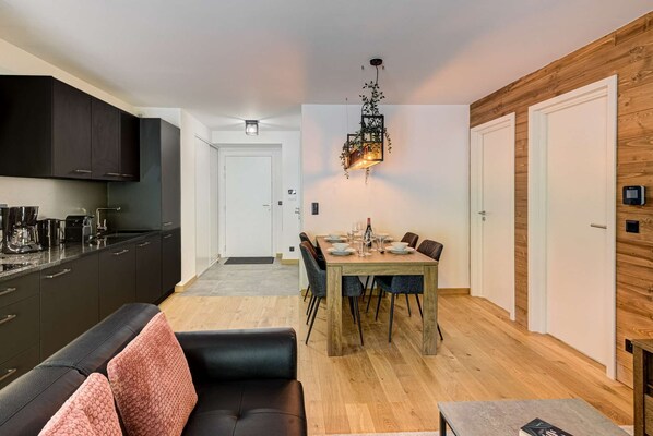 Una cocina moderna con armarios oscuros, una zona de comedor con una mesa y sillas de madera, y una acogedora sala de estar con un sofá negro.