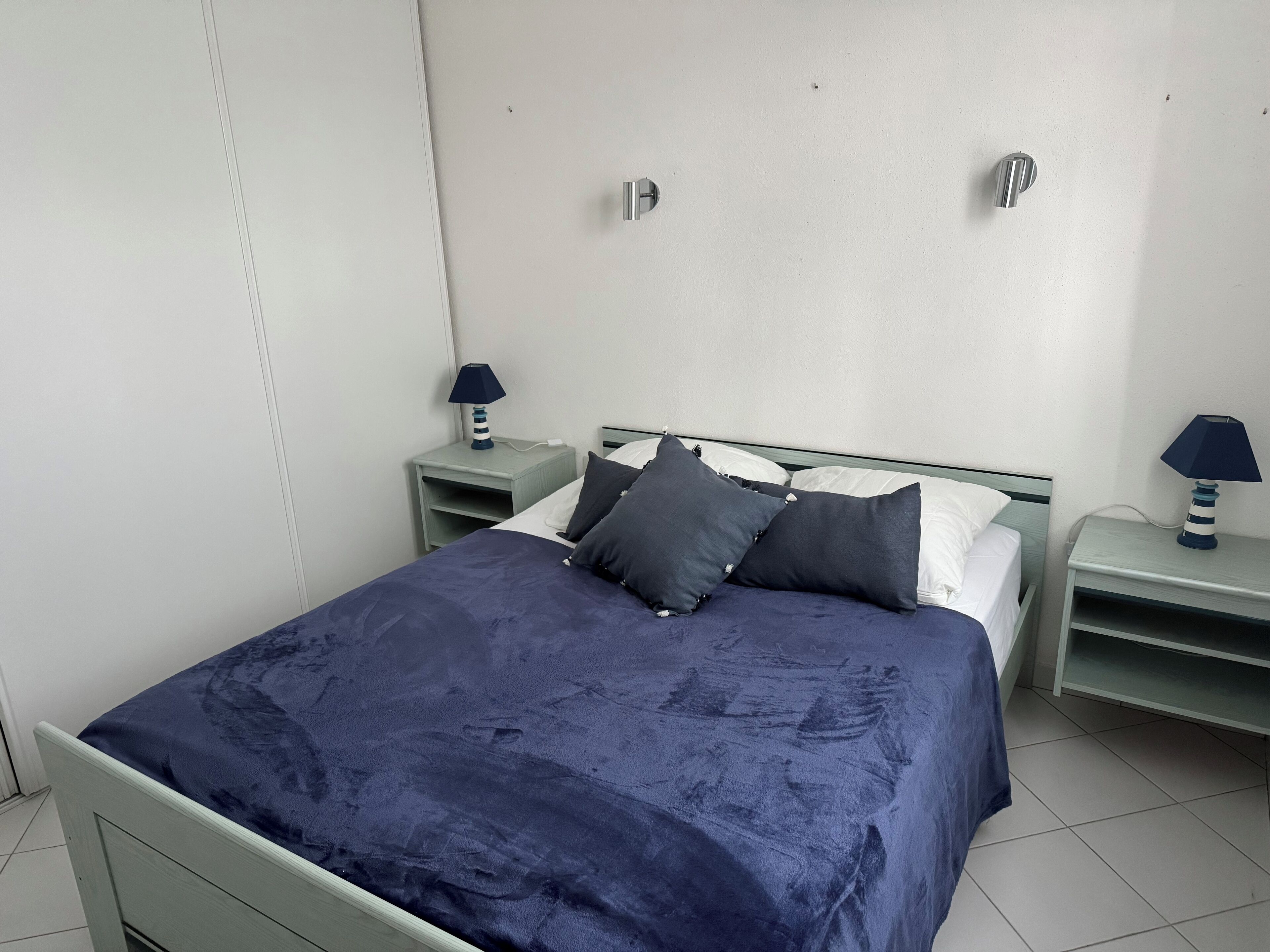 A neatly made bed with a dark blue bedspread and pillows, two bedside tables with lamps, and a white wall with a door.