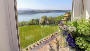 A view from a window overlooking a lake, a grassy lawn, and a stone pathway leading to a building. - Menai View (Menai Bridge)
