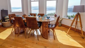 A dining area with a wooden table and chairs, large windows with views of the sea, and a tripod lamp. - Menai View (Menai Bridge)