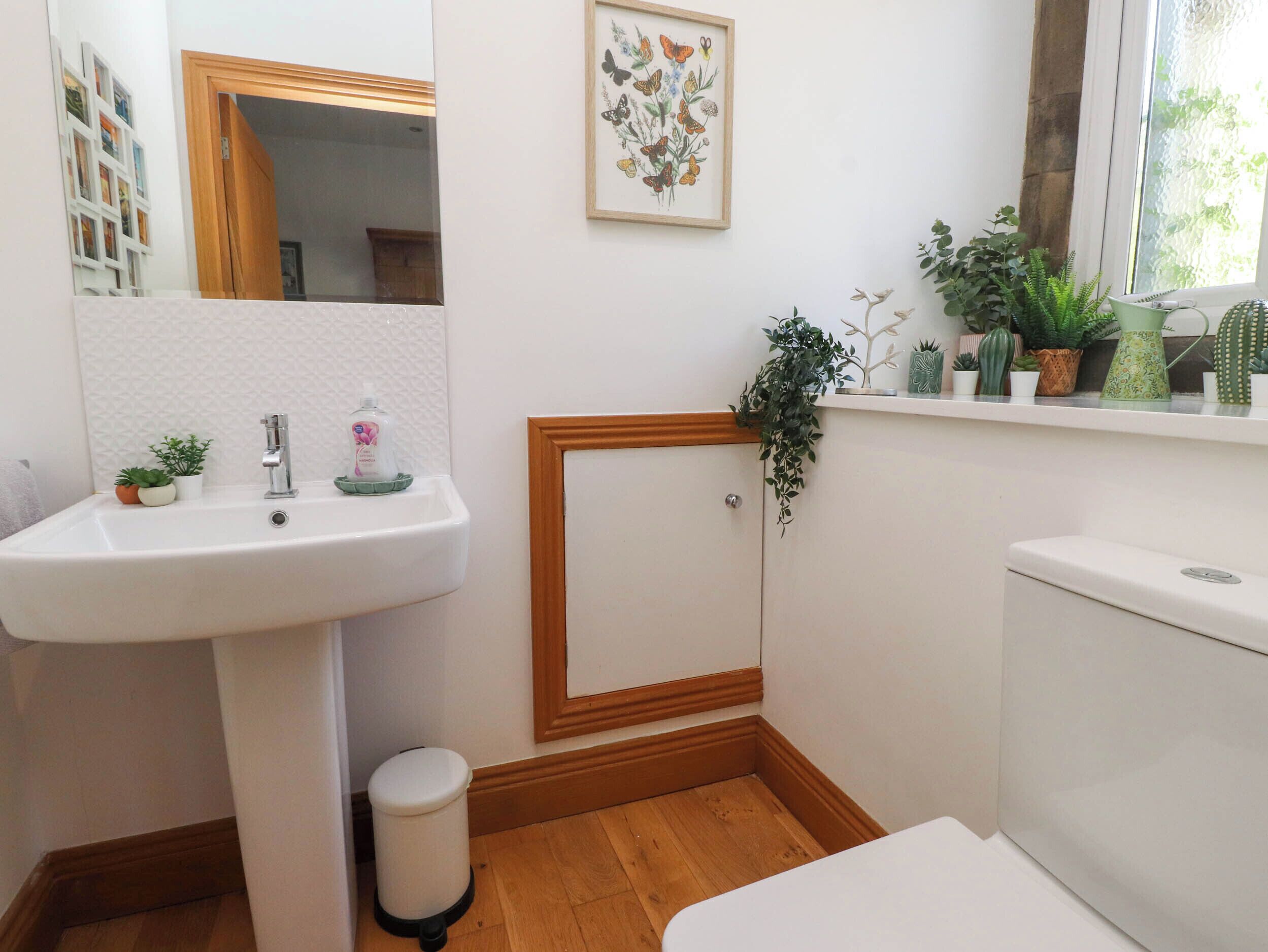 A bathroom with a white sink, mirror, and toilet. There are potted plants on the windowsill and a framed botanical print on the wall.