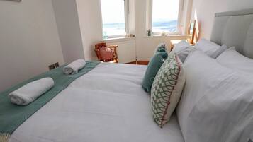 Una cama bien tendida con sábanas blancas y un plaid verde, una silla de madera con un cojín rosa, y vistas al exterior a través de una ventana.