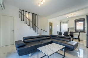 Un salon moderne avec un canapé d’angle en cuir noir, une table basse en marbre, un escalier avec des rampes noires et, au fond, un espace cuisine.