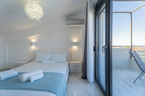 A modern bedroom with a large bed, bedside tables, an air conditioning unit, and a balcony with a chair.