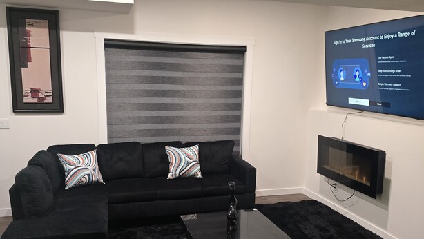 A modern living room with a black sofa, a black coffee table, and a large flat-screen TV mounted on the wall. - Cozy 2-Bedroom Suite: 5 Minutes Walk To Chestermere Lake (Chestermere)