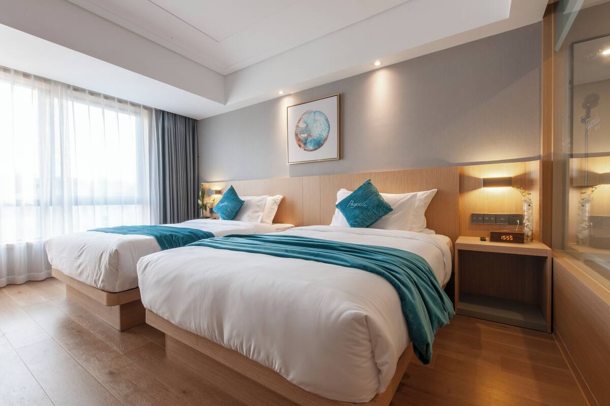 Una habitación de hotel moderna con dos camas, un ventanal con cortinas translúcidas, una obra de arte colgada en la pared y una mesita de noche con un reloj.