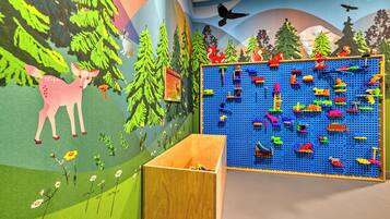Zona de juegos infantiles con un gran panel de buratos azul lleno de juguetes coloridos, un mural con paisaje de bosque y una estructura de madera para jugar.
