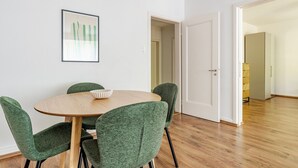 Un coin repas avec une table ronde, quatre chaises à revêtement vert, un bol blanc et un tableau encadré accroché au mur.