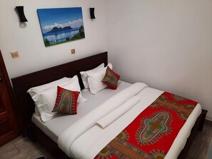 A neatly made bed with white linens and two decorative pillows, a red and green patterned bedspread, and a painting of a lake and mountains on the wall.