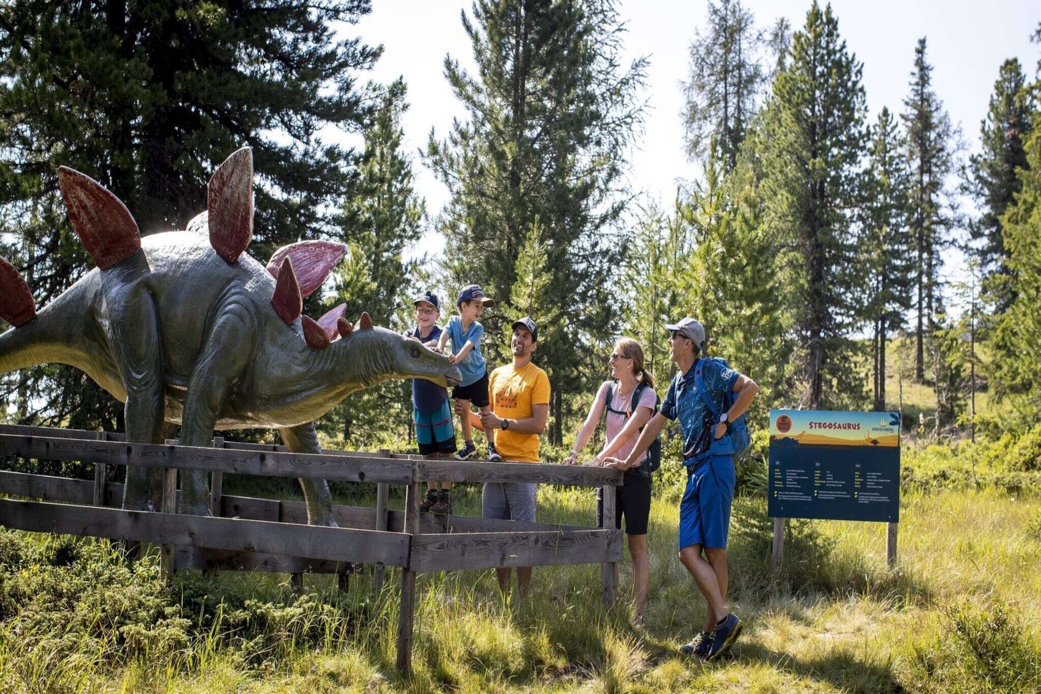 A group of people observing a dinosaur exhibit in a natural setting with a wooden fence and a signboard.