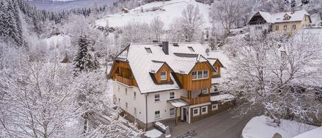 Un edificio de madera de varios pisos con techo nevado, rodeado de árboles cubiertos de nieve y un paisaje montañoso al fondo.