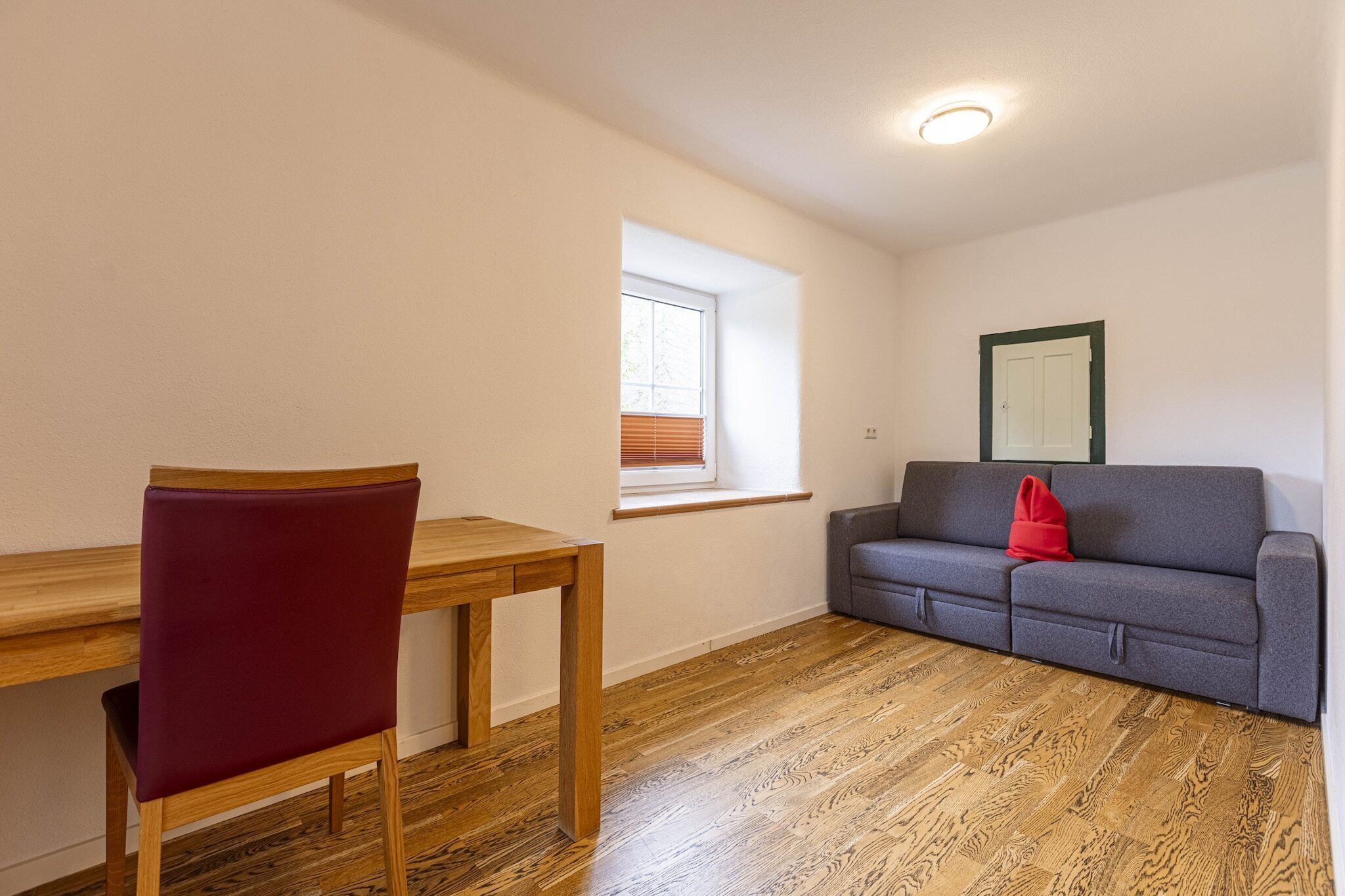 Una habitación con piso de madera, una mesa de comedor con una silla, un sofá gris con un cojín rojo y una ventana con persianas.