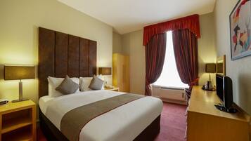 A hotel room with a large bed, a desk with a computer, a wardrobe, and a window with curtains.