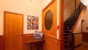 Un pasillo con puerta de madera, un cartel en la pared con números de habitaciones, una escalera con barandilla negra y una mesa con revisteros.