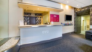 A reception desk with a digital clock, a sign with the logo 'OYO', and a flat-screen TV mounted on the wall.