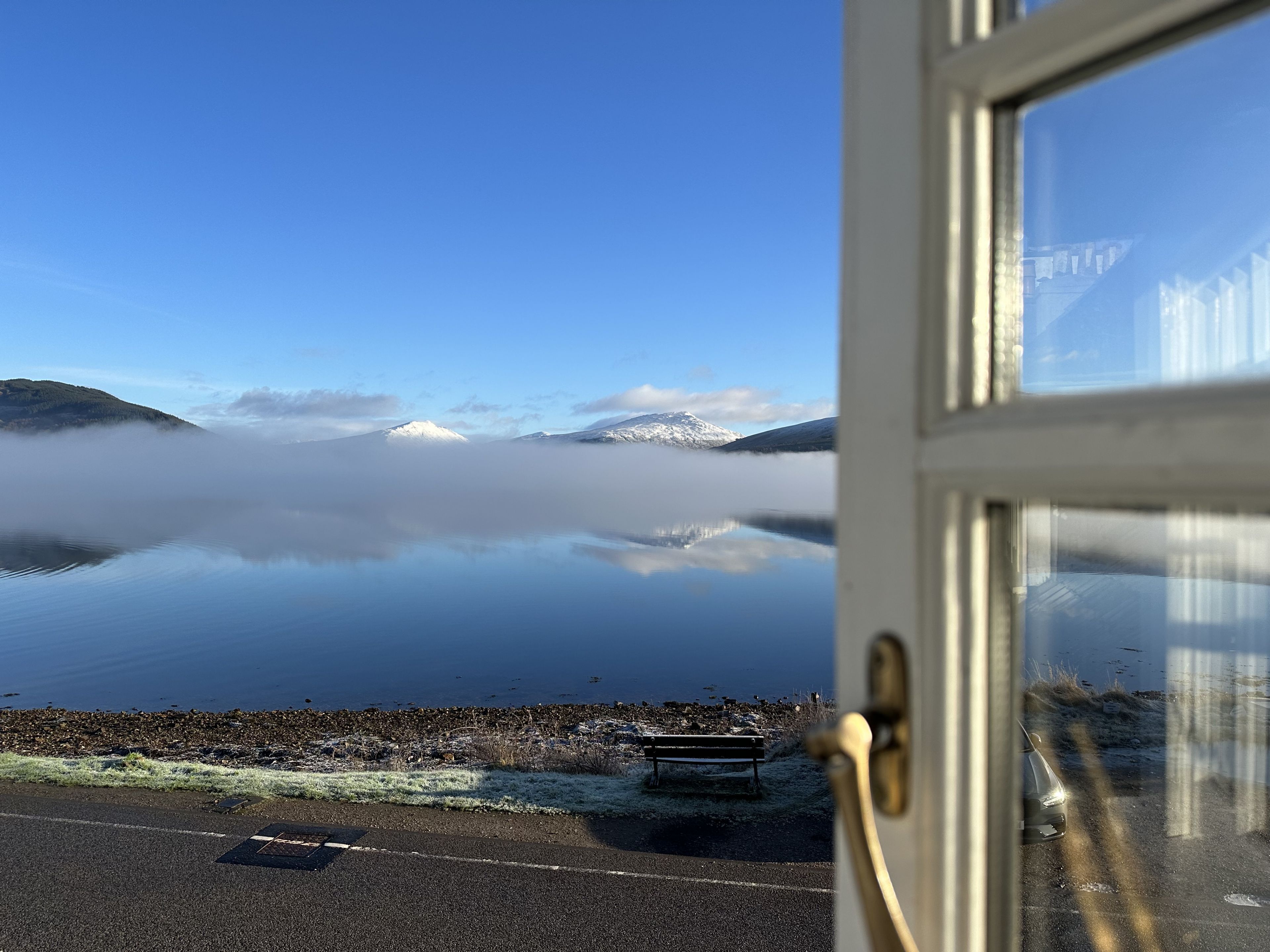 Vista a través de una ventana que muestra un lago con montañas al fondo, un banco y un coche aparcado en la orilla.
