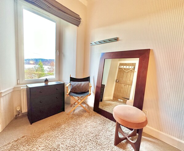 Un dormitorio con un ventanal grande, un aparador, una silla plegable, un taburete y un espejo.