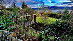 A wooden bridge over a stream, surrounded by greenery and a view of hills and a lake.
