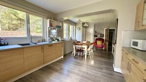 A modern kitchen with wooden cabinets, a dining area with a table and chairs, and a view of the outdoors through large windows.