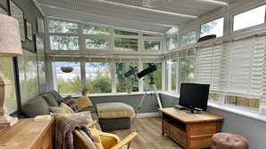 A sunroom with a wooden desk, a comfortable seating area, a telescope, and a television.