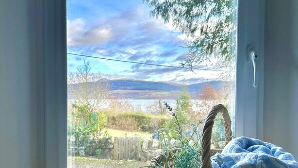 A window with a view of a landscape, a wicker basket with a blue cushion, and a clock on a shelf.
