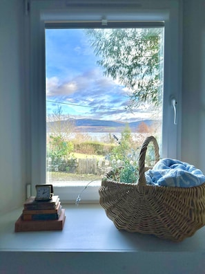 Una ventana con vistas al paisaje, una cesta de mimbre con un cojín azul y un reloj sobre una estantería.