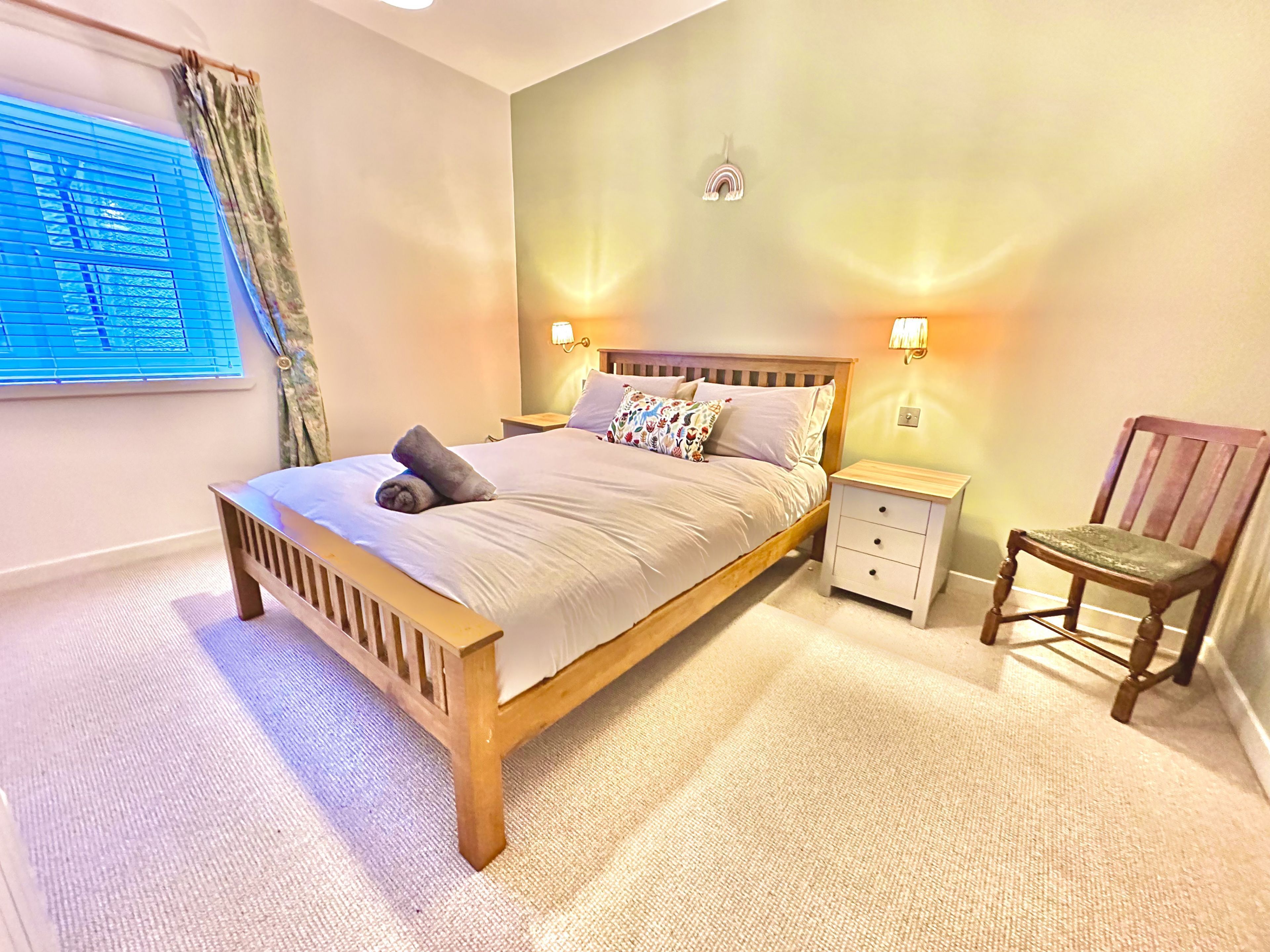 Quarto com cama de madeira, mesas de cabeceira, uma cadeira, janela com persianas e luminária fixada na parede.