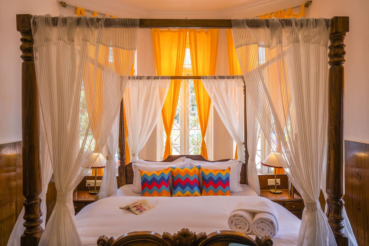 A four-poster canopy bed with a white bedspread and colorful zigzag pillows, a wooden nightstand with a lamp, and a window with yellow curtains.