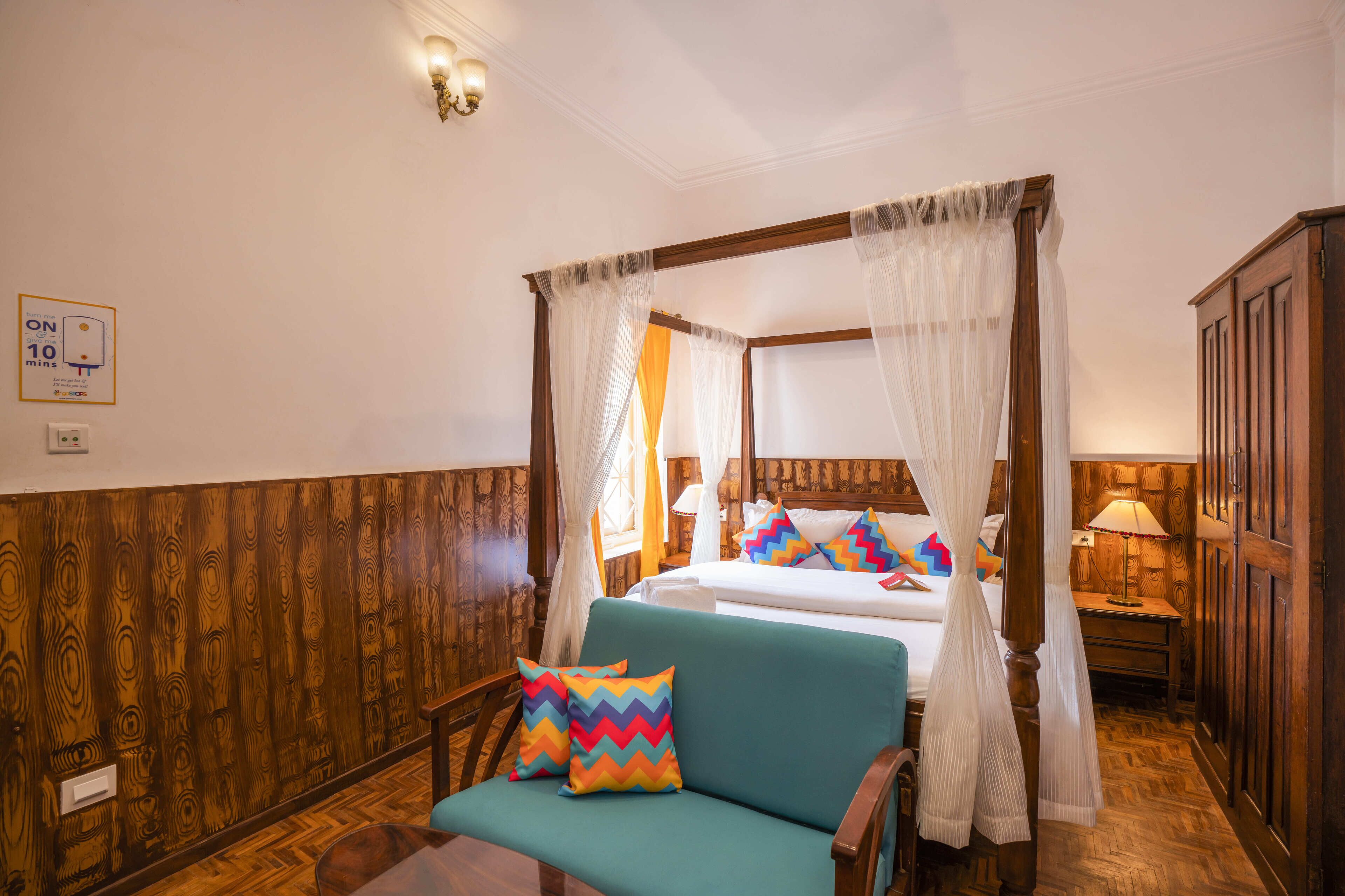 A four-poster bed with white curtains, a teal sofa with colorful pillows, and a wooden floor.