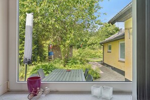 A window view of a garden with a wooden table and chairs, a pink bucket, and a bicycle.