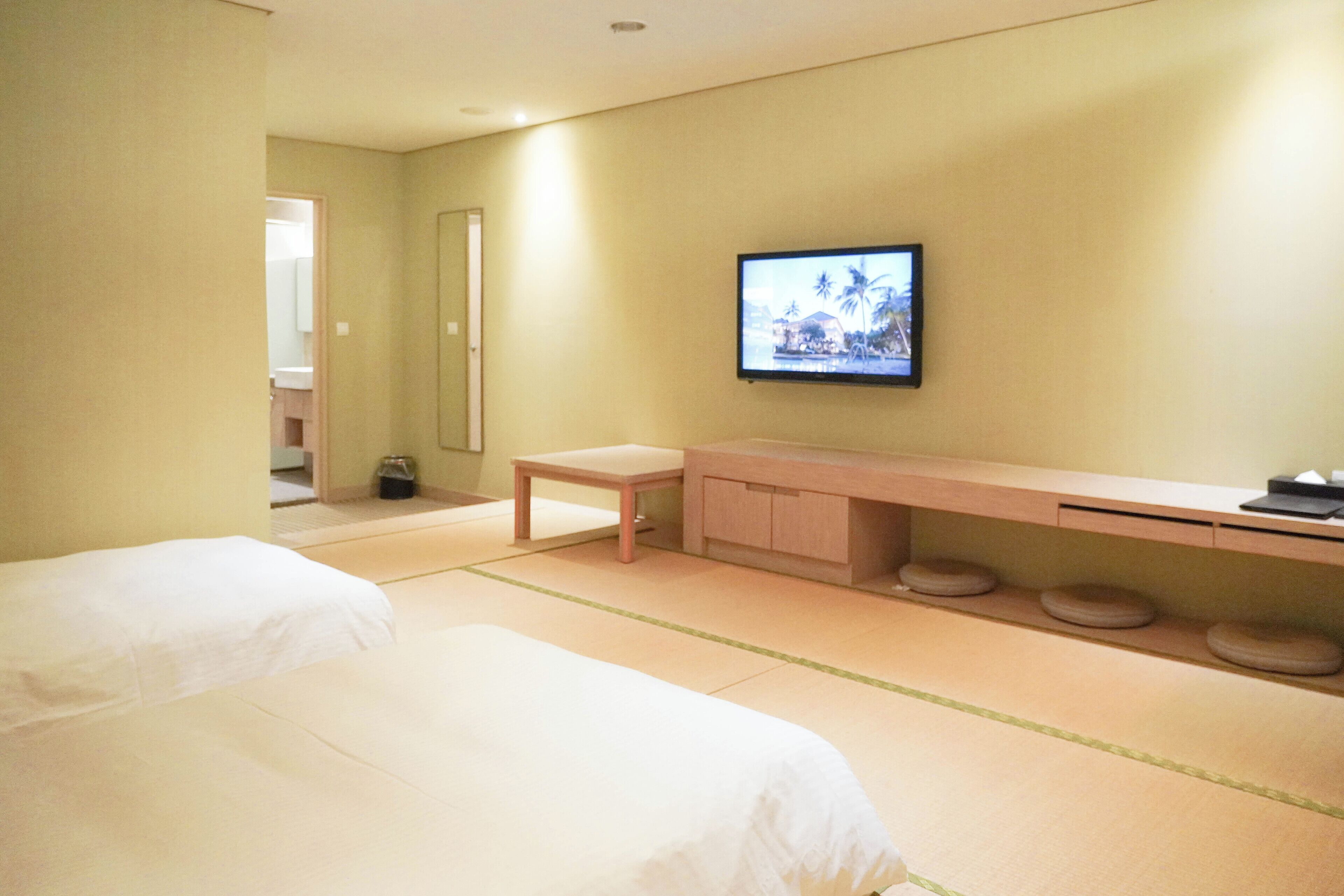 A hotel room with two beds, a wooden bench, a TV mounted on the wall, and a small table.