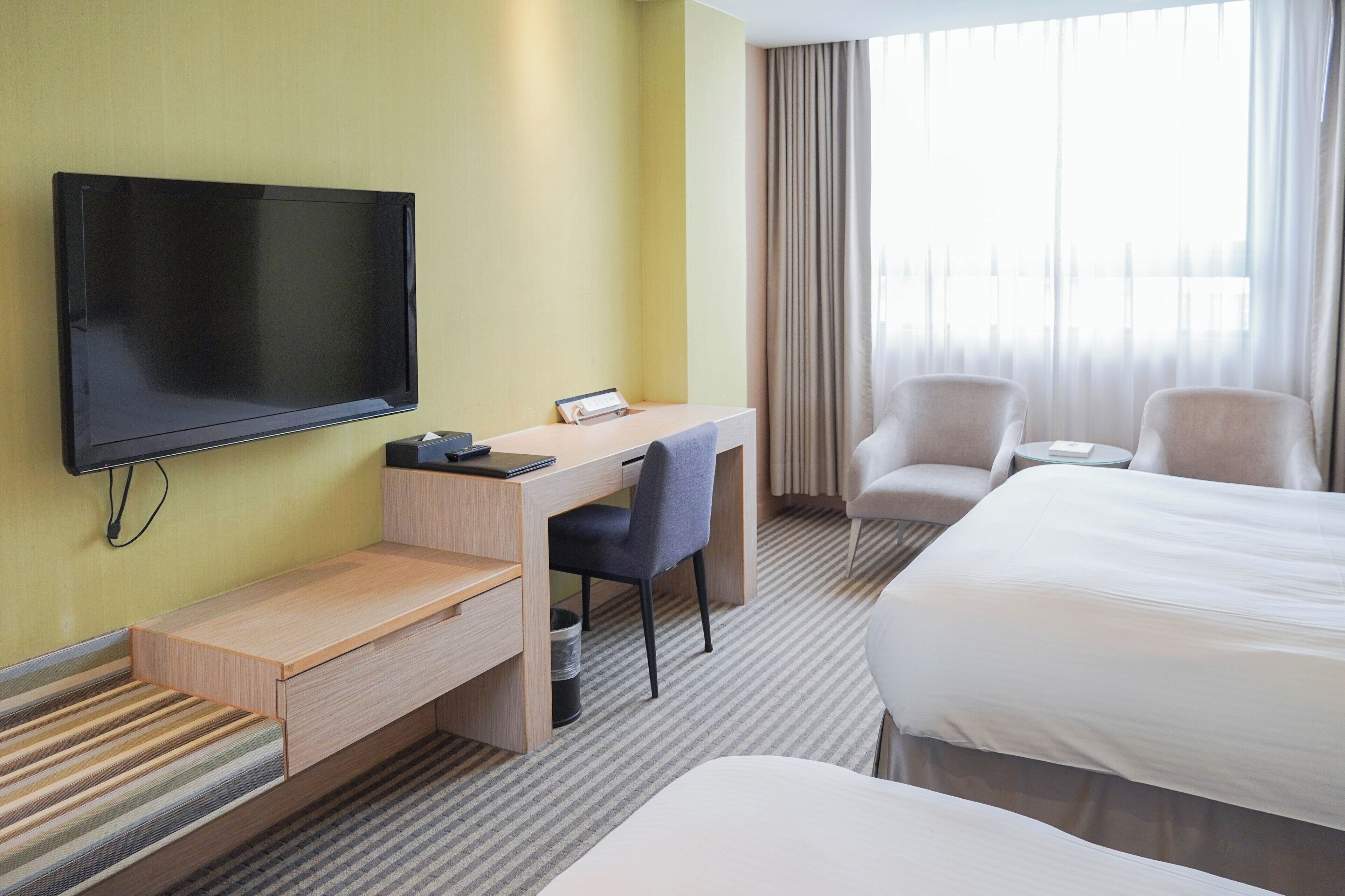Une chambre d’hôtel équipée d’un téléviseur à écran plat, d’un bureau avec une chaise, de deux fauteuils, d’un lit et d’une grande fenêtre avec des rideaux.